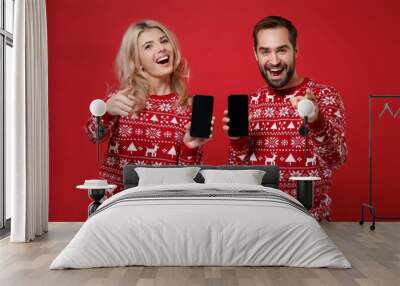 Merry young couple guy girl in Christmas sweaters posing isolated on red background. New Year 2020 celebration concept. Mock up copy space. Hold mobile phone with blank screen point fingers on camera. Wall mural