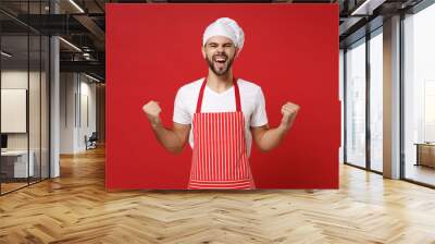 Overjoyed bearded male chef cook or baker man in striped apron white t-shirt toque chefs hat posing isolated on red background. Cooking food concept. Mock up copy space. Scream, doing winner gesture. Wall mural