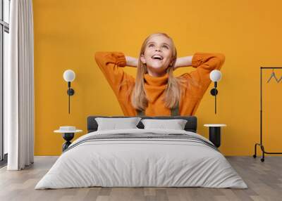 Pensive little blonde kid girl 12-13 years old in turtleneck, jumpsuit isolated on orange yellow wall background. Childhood lifestyle concept. Mock up copy space. Looking up with hands behind head. Wall mural