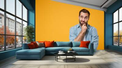 Pensive young bearded man in casual blue shirt posing isolated on yellow orange background studio portrait. People emotions lifestyle concept. Mock up copy space. Put hand prop up on chin, looking up. Wall mural