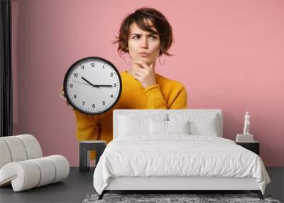 Pensive young brunette woman girl in yellow sweater posing isolated on pastel pink background, studio portrait. People lifestyle concept. Mock up copy space. Holding clock, put hand prop up on chin. Wall mural