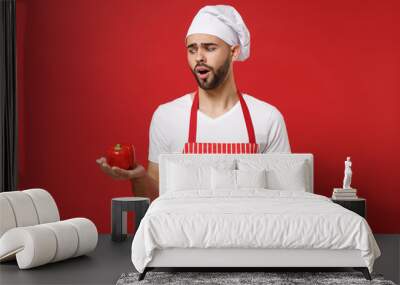 Perplexed young male chef cook or baker man in striped apron white t-shirt toque chefs hat isolated on red background. Cooking food concept. Mock up copy space. Holding fresh vegetable bell pepper. Wall mural