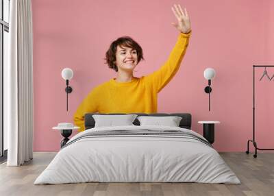 Pleasant young brunette woman girl in yellow sweater posing isolated on pink background studio portrait. People lifestyle concept. Mock up copy space. Waving and greeting with hand as notices someone. Wall mural