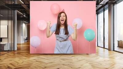 Portrait of attractive joyful young woman in birthday hat blue dress doing winner gesture screming on pink background with colorful air baloons. Birthday holiday party, people sincere emotion concept. Wall mural