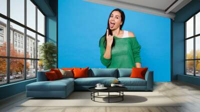 Portrait of cheerful young woman in green casual clothes showing tongue depicting heavy metal rock sign isolated on bright blue wall background in studio. People lifestyle concept. Mock up copy space. Wall mural