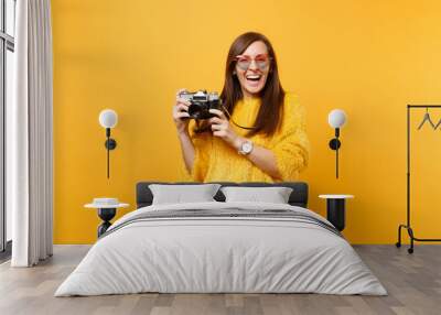 Portrait of laughing young woman in heart eyeglasses taking pictures on retro vintage photo camera isolated on bright yellow background. People sincere emotions, lifestyle concept. Advertising area. Wall mural