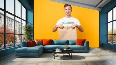 Portrait of man in white t-shirt written inscription green title volunteer hold blank craft paper bag for takeaway isolated on yellow background. Voluntary free assistance help charity grace concept. Wall mural