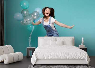 Portrait of pretty funny young woman in denim clothes jumping high celebrating and holding colorful air balloons isolated on blue turquoise background. Birthday holiday party, people emotions concept. Wall mural