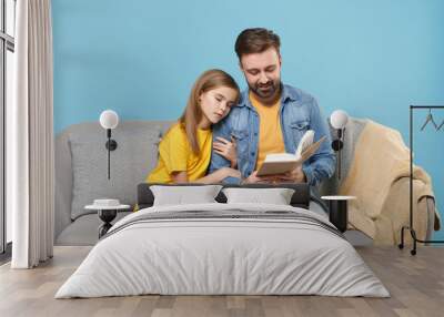 Pretty bearded man in casual clothes with cute child baby girl. Father little daughter isolated on pastel blue background. Love family parenthood childhood concept. Sit on couch reading book hugging. Wall mural