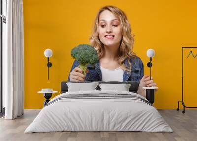 Pretty young woman in denim clothes isolated on yellow orange background. Proper nutrition vegetarian food, healthy lifestyle diet meal. Diabetes dieting concept. Hold green broccoli, biting lips. Wall mural