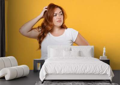 Puzzled pensive young redhead plus size body positive female woman 20s in white blank design casual t-shirt posing put hand on head looking aside isolated on yellow color background studio portrait. Wall mural