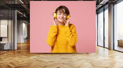 Relaxed young brunette woman girl in yellow sweater posing isolated on pastel pink background in studio. People lifestyle concept. Mock up copy space. Listen music with headphones keeping eyes closed. Wall mural