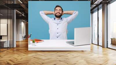 Relaxed young man in light shirt sit work at desk with pc laptop isolated on pastel blue background. Achievement business career lifestyle concept. Keeping eyes closed sleeping with hands behind head. Wall mural