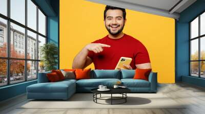 Satisfied smiling cool cheerful fun young happy Indian man wears red t-shirt casual clothes hold in hand use point index finger on mobile cell phone isolated on plain yellow orange background studio. Wall mural