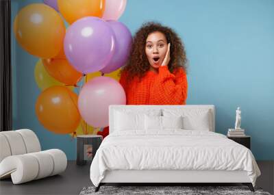 Shocked african american girl in orange knitted clothes isolated on pastel blue background. Birthday holiday party people emotions concept. Celebrating holding colorful air balloons put hand on cheek. Wall mural