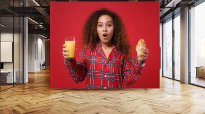 Shocked african american girl in pajamas homewear resting at home isolated on red background. Relax good mood lifestyle concept. Mock up copy space. Eating breakfast, hold croissant, glass of juice. Wall mural