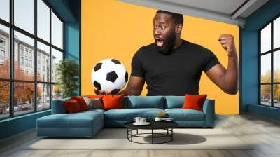 Shocked african american man guy football fan in black t-shirt isolated on yellow background. Sport family leisure lifestyle concept. Cheer up support favorite team with soccer ball, clenching fist. Wall mural