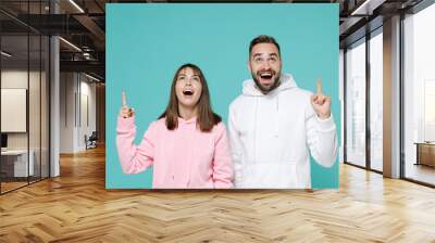 Shocked amazed surprised young couple two friends man woman 20s wearing white pink casual hoodie standing pointing index fingers up isolated on blue turquoise colour wall background studio portrait. Wall mural