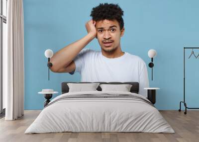 Shocked perpelxed young african american guy in casual white t-shirt posing isolated on blue background studio portrait. People sincere emotion lifestyle concept. Mock up copy space. Put hand on head. Wall mural