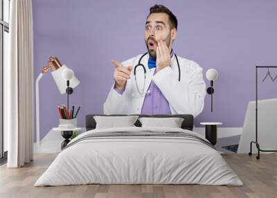 Shocked surprised male doctor man in white medical suit sit at desk work on computer documents in clinic office point point inddex finger aside hold face isolated on violet background studio portrait Wall mural