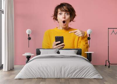 Shocked young brunette woman girl in yellow sweater posing isolated on pastel pink wall background studio portait. People lifestyle concept. Mock up copy space. Pointing index finger on mobile phone. Wall mural