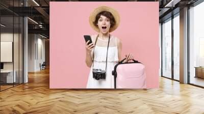 Shocked young tourist girl in summer dress hat with photo camera isolated on pink background. Traveling abroad to travel weekend getaway. Air flight journey concept. Using mobile phone, hold suitcase. Wall mural