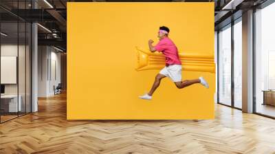 Side view excited happy young man he wearing pink t-shirt near hotel pool jump high run fast hurrying up hold inflatable mattress isolated on plain yellow background. Summer vacation sea rest concept. Wall mural