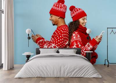 Side view merry young couple two man woman wear red Christmas sweater Santa hat posing hold use mobile cell phone isolated on plain pastel light blue background. Happy New Year 2023 holiday concept. Wall mural