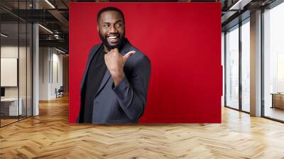 Side view of cheerful funny young african american business man 20s wearing classic jacket suit standing pointing thumb aside on mock up copy space isolated on bright red background studio portrait. Wall mural