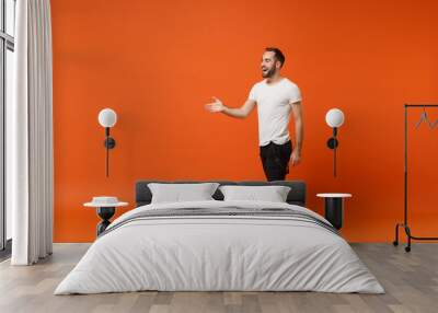 Side view of cheerful young man in casual white t-shirt posing isolated on orange wall background in studio. People lifestyle concept. Mock up copy space. Standing with outstretched hand for greeting. Wall mural