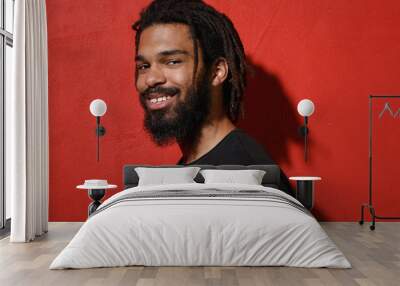 Side view of handsome smiling young african american man guy with dreadlocks 20s wearing black casual t-shirt posing looking camera isolated on bright red color wall background studio portrait. Wall mural