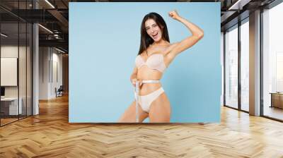 Side view of happy young brunette woman 20s in beige brassiere underwear standing posing measuring waist with measure tape doing winner gesture isolated on pastel blue background studio portrait. Wall mural