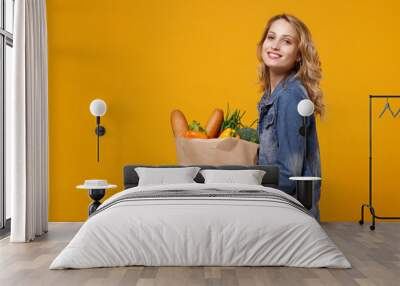 Side view of smiiling woman in denim clothes isolated on orange background. Delivery service from shop or restaurant concept. Hold brown craft paper bag for takeaway with food products looking camera. Wall mural