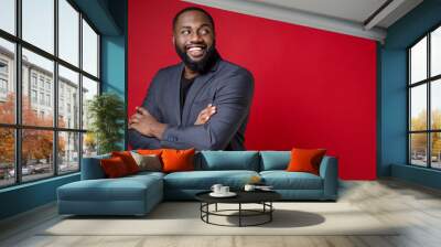 Side view of smiling attractive young african american business man 20s in classic jacket suit standing holding hands crossed looking aside isolated on bright red color background studio portrait. Wall mural