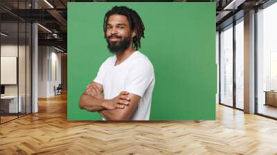 Side view of smiling funny young african american man guy 20s wearing white casual t-shirt posing holding hands folded crossed looking camera isolated on green color wall background studio portrait. Wall mural