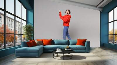 Side view of surprised young brunette woman girl in casual red hoodie blue jeans posing isolated on grey background in studio. People lifestyle concept. Mock up copy space. Point index fingers aside. Wall mural