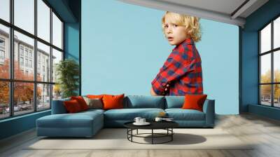 Side view of upsey offended little curly kid boy 10s years old wearing basic red checkered shirt looking camera isolated on blue color background children studio portrait. Childhood lifestyle concept. Wall mural