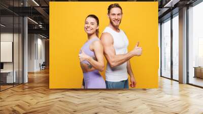 Side view young fitness trainer instructor sporty two man woman wear blue clothes spend time in home gym stand back to back show thumb up isolated on plain yellow background Workout sport fit concept Wall mural