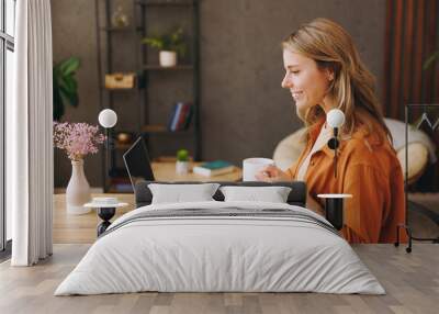 Side view young IT woman wearing orange shirt hold use laptop pc computer drink coffee sit alone at table in coffee shop cafe restaurant indoors work or study Freelance mobile office business concept Wall mural