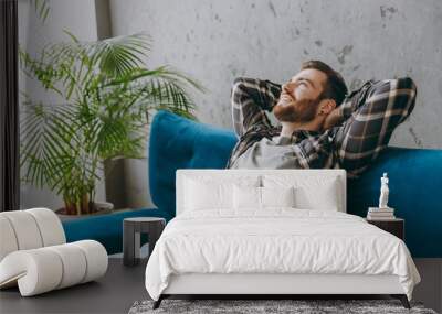 Side view young minded pensive dreamful happy man 20s he wear shirt hold hands behind neck sitting on blue sofa in own living room apartment stay home indoor flat on weekends People lifestyle concept Wall mural