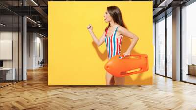 Side view young sexy lifeguard woman slim body wear striped red blue one-piece swimsuit hold aid lifebuoy isolated on vivid yellow color wall background studio. Summer hotel pool sea rest sun concept. Wall mural