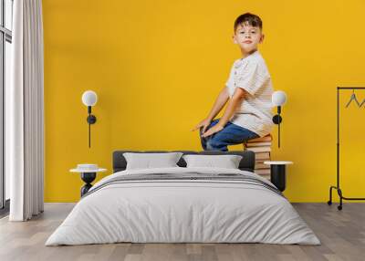 Side view young smart wunderkind school boy 5-6 years old in t-shirt casual clothes sit on pile of books isolated on plain yellow background studio Childhood children kids education lifestyle concept. Wall mural