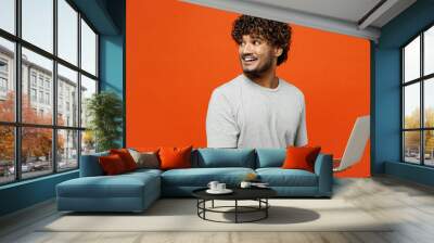 Side view young smiling happy Indian man wears t-shirt casual clothes hold use work on laptop pc computer look aside on area isolated on orange red color background studio portrait. Lifestyle concept. Wall mural