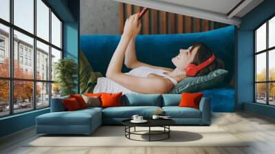 Side view young smiling happy woman wear white tank shirt headphones listen music use mobile cell phone lay down on blue sofa stay at home flat rest relax spend free spare time in living room indoor. Wall mural