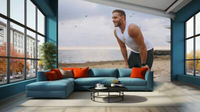 Side view young strong sporty athletic toned fit sportsman man he wear sports clothes lean on knees warm up training at sunrise sun dawn over sea beach outdoor on pier seaside in summer day morning. Wall mural