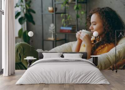 Sideways young woman of African American ethnicity wear t-shirt drink coffee read book close eyes sits in armchair stay at home flat rest relax spend free spare time in living room indoor grey wall. Wall mural