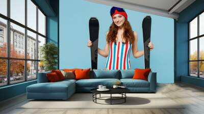 Skier smiling happy cheerful confident fun woman 20s wear warm striped swimsuit goggles mask spend extreme weekend in mountains resort holding ski look camera isolated on plain blue background studio Wall mural