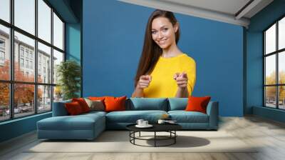 Smiling cheerful attractive beautiful young brunette woman 20s wearing yellow casual clothes posing standing pointing index fingers on camera isolated on blue color wall background studio portrait. Wall mural