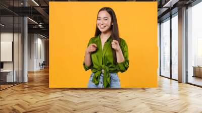 Smiling cheerful beautiful attractive young brunette asian woman 20s wearing basic green shirt standing pointing index fingers on camera isolated on bright yellow colour background studio portrait. Wall mural