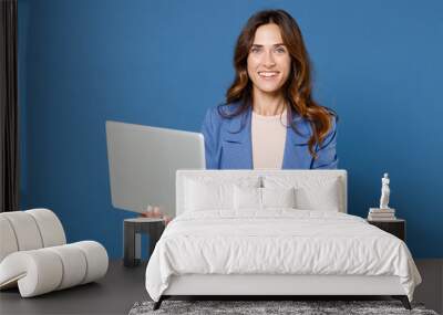 Smiling cheerful beautiful attractive young brunette woman 20s wearing basic jacket standing working on laptop pc computer looking camera isolated on bright blue colour background studio portrait. Wall mural